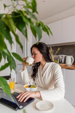 Rahat beyaz cüppeli genç bir kadın mutfaktaki masada otururken dizüstü bilgisayarla çalışıyor. Kahvaltıda evde çalış. İş kadını sosyal mesafeyi koruyor, uzaktan çalışıyor. Yeni normal kavram