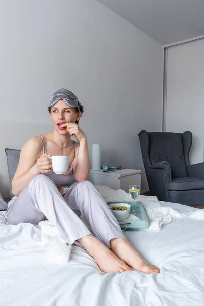Young woman in silk pajamas has healthy breakfast while lying on bed in bedroom. Home wellbeing concept. Emotional health of a young woman