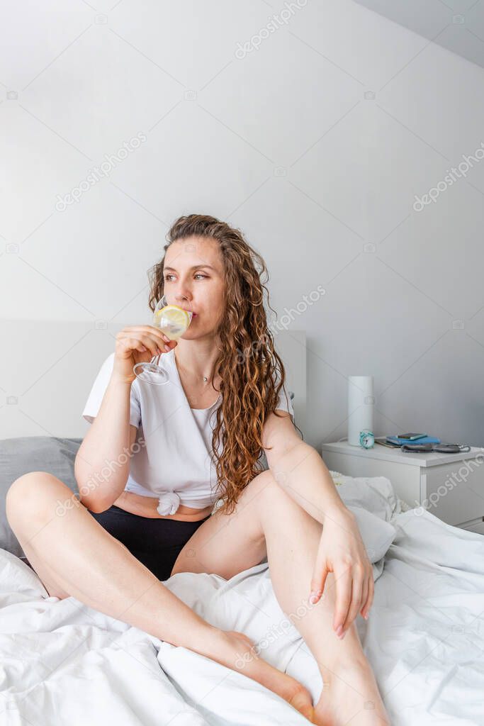 Young woman ginger hair sitting the bed in bedroom and drinks water with lemon. Fresh morning. Wellness and detox concept. Copy space