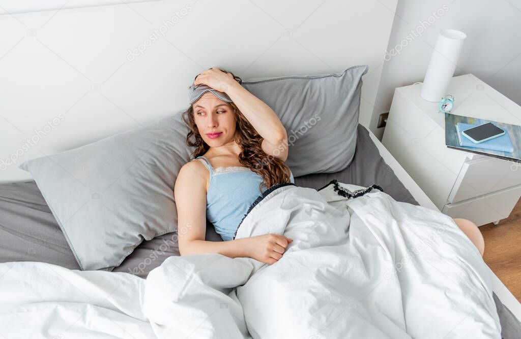 Young woman in silk pajama yawns while sitting in comfortable soft bed. Home well being. Wellness concept. Copy space