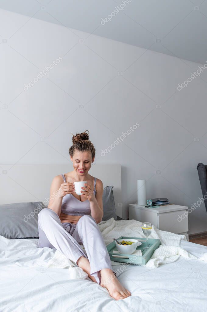 Young woman in silk pajamas has healthy breakfast while lying on bed in bedroom. Home wellbeing concept. Emotional health of a young woman