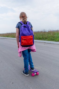 Kot pantolonlu ve pembe kazaklı genç bir kız sokakta kaykaya biniyor. Genç yaşam tarzı konsepti. Uzayı kopyala Arkasında mor bir sırt çantası var..