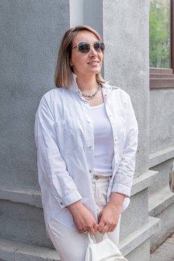 Genç bir kadın sırt çantasını elinde tutuyor ve gri binanın yanında duruyor. Beyaz saçlı kız beyaz takım elbise giyiyor. İş konsepti. Şehir manzarası