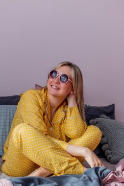 Yatakta sarı pijamalarıyla fotoğrafçıya poz veren genç bir kadın. Teknoloji kavramı. Duygular yaşam tarzı