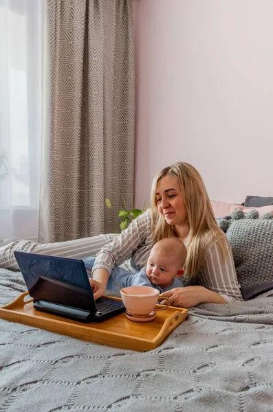 Young woman in comfortable home clothes work at laptop with a baby lying on bed in bedroom. Home wellbeing concept. Emotional health a mom woman