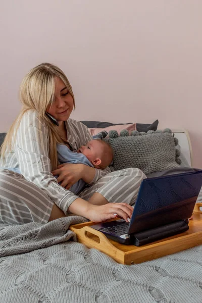 Young woman in comfortable home clothes works on laptop with a baby in her arms in bedroom. Home wellbeing concept. Emotional health mom woman
