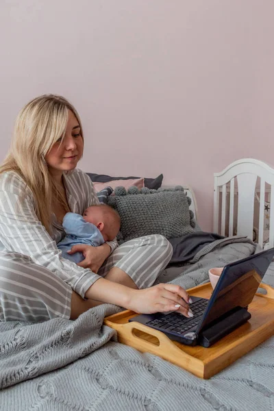 Young woman in comfortable home clothes works on laptop with a baby in her arms in bedroom. Home wellbeing concept. Emotional health mom woman