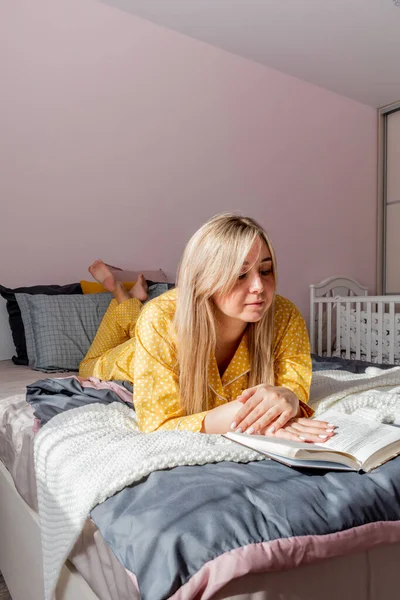 Young woman in yellow pajamas read book while lying on bed in bedroom. Home wellbeing concept. Emotional health of a young woman