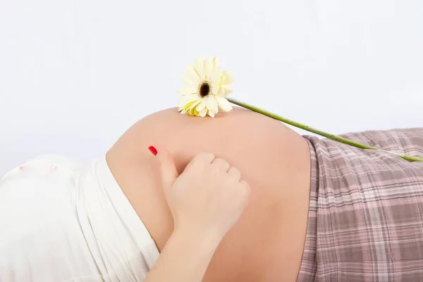 Mulher grávida deita-se na cama em um fundo branco e mostra os polegares para cima. flor amarela na barriga — Fotografia de Stock