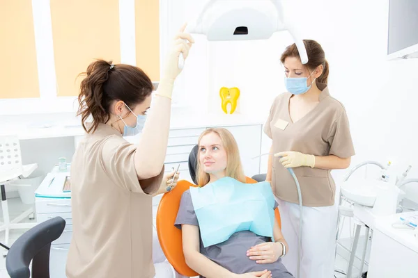 Young pregnant woman at the dental clinic, sitting in the treatment chair, the dentist and her assistant are preparing to treat the patient — стоковое фото