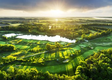 Golf sahası manzarası, yeşil çim yolları ve ormanla dramatik gündoğumunda.