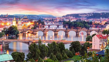 Çek Cumhuriyeti Prag 'daki Vltava Nehri üzerindeki aydınlık köprülerin akşam manzarası, tarihi kuleler, şehir ışıkları ve romantik atmosfer yaratan yansımalar.