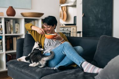 Kanepede köpeğiyle oturan ve abur cubur yiyen kadın.