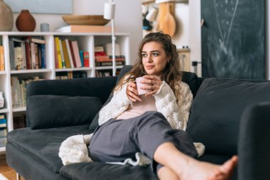 Kadın, evindeki kanepede otururken bir fincan kahvenin tadını çıkarıyor.