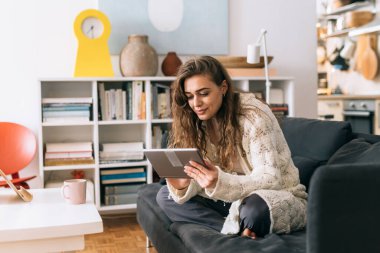 Kadın evindeki kanepede oturup, sabah kahvesini içip, dijital tablet kullanıyor.