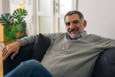 Olumlu gülümseyen sakallı adam evindeki koltukta oturuyor ve kameraya bakıyor.