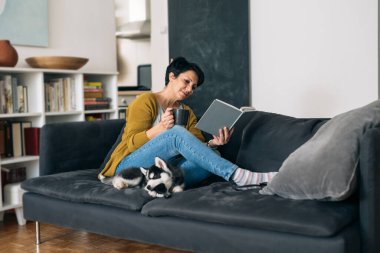 Oturma odasındaki kanepede oturmuş kitap okuyan bir kadın.