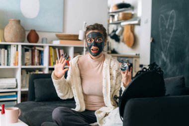 Kadın oturma odasında otururken kozmetik kilden maske takıyor.