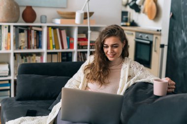 Kadın evindeki kanepede dinlenirken dizüstü bilgisayar kullanıyor ve sabah kahvesini içiyor.