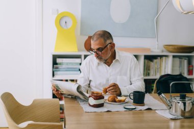 Orta yaşlı bir adam evde kahvaltı yapıyor. Gazete okuyor.