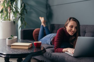 Kadın kanepeye uzanmış ve evinde dizüstü bilgisayar kullanıyor.