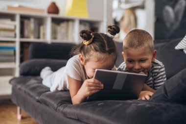 Çocuklar evde birlikte oynuyorlar. Kanepede yatarken dijital tablet kullanıyorlar..