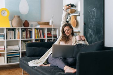 Kadın kanepede dinleniyor, dizüstü bilgisayar kullanıyor ve evde kahve içiyor.