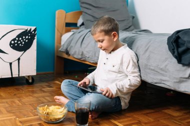 Küçük çocuk sağlıksız yiyecekler yiyor ve dijital tablet ya da dizüstü bilgisayar kullanıyor. Evindeki odasında oturuyor.