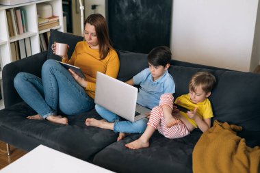 Asosyal aile. Anne ve iki çocuğu kanepede oturup dijital cihazlar kullanıyorlar.