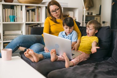Anne ve çocukları evdeki kanepede otururken dizüstü bilgisayar kullanıyorlar.