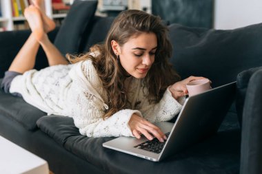 Kadın kanepede dinleniyor ve evde dizüstü bilgisayar kullanıyor.