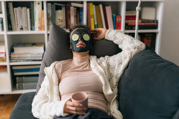 woman with cosmetic face mask applied on her face relaxing on sofa at her home