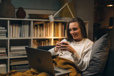 Kadın evdeki kanepede dinleniyor ve dizüstü bilgisayar kullanıyor.