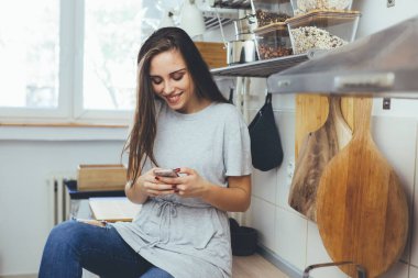 Genç bir kadın mutfak tezgahında otururken akıllı telefon kullanıyor.