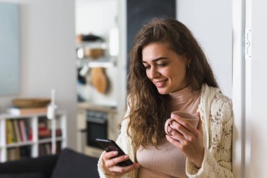 Kadın cep telefonu kullanıyor ve sabah kahvesini evde içiyor.