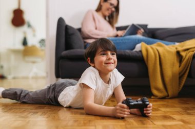 Çocuk evde video oyunu oynuyor. Annem arka planda bulanık bir şekilde kanepede oturuyor.