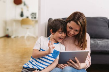 Anne ve oğlu evde oturup dijital tablet kullanıyorlar. Video görüşmeleri var.