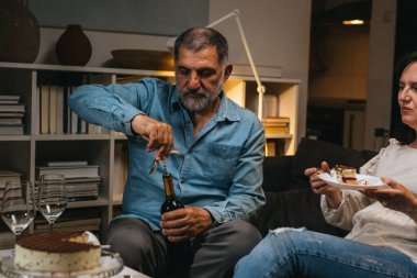 Çift evde yıldönümünü kutluyor. Adam şarap şişesini açıyor.
