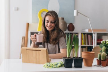 Kadın odasında oturmuş dijital tablet kullanıyor ve kahve ya da çay içiyor.