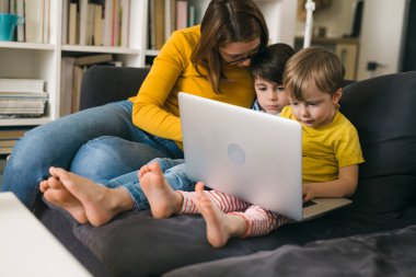 Anne ve çocukları dizüstü bilgisayar kullanırken, evde bir kanepede oturuyor.