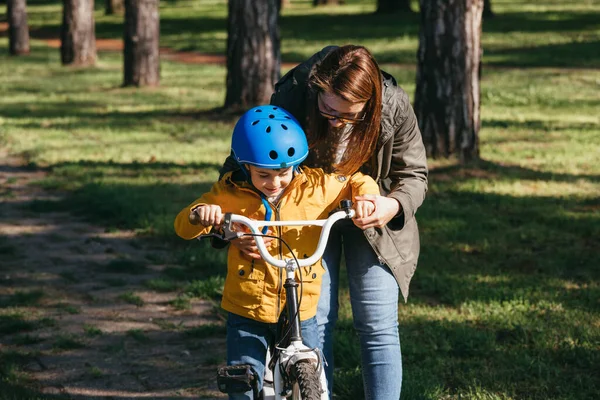 Küçük çocuk annesiyle birlikte parkta bisiklet sürmeyi öğreniyor.