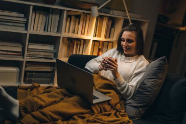 Kanepede oturan ve evinde dizüstü bilgisayar kullanan genç bir kadın. Kahve ya da çay içiyor.