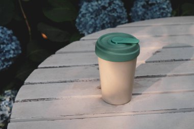 Çiçek açan yaz arkaplanına karşı beyaz ahşap bahçe masasının kapağı ile yeniden kullanılabilir çevre dostu bambu kupası. Uzayı kopyala Sıfır atık, sürdürülebilir yaşam tarzı konsepti. Plastik atıkları azalt.