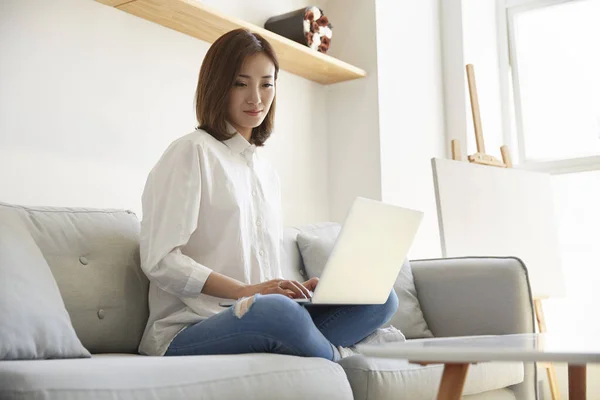 自宅のソファでノートパソコンを使って仕事をしている若いアジア人のビジネス女性は — ストック写真