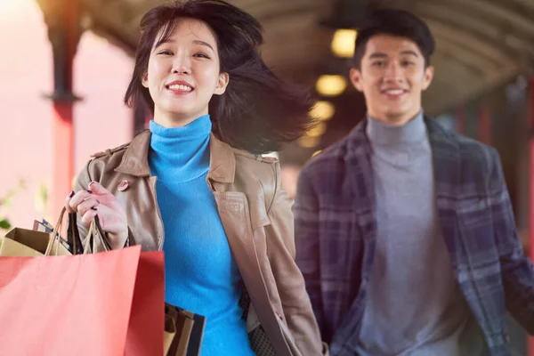 若いアジア人のカップルが歩道を走り買い物袋を手に笑顔で走り — ストック写真