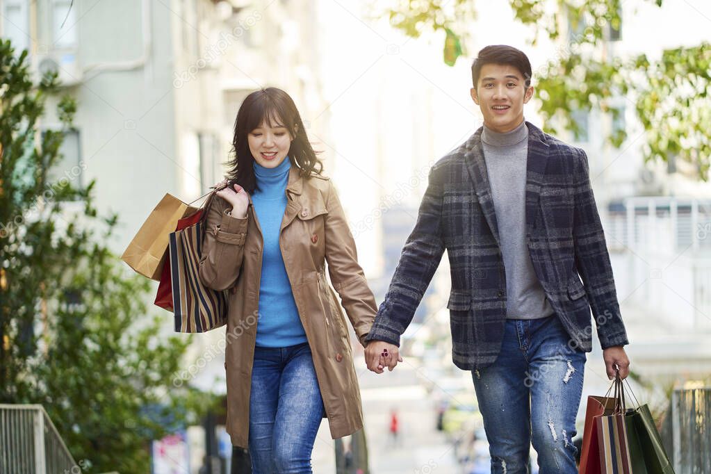 happy young asian couple walking on street in the city with shopping bags in hands