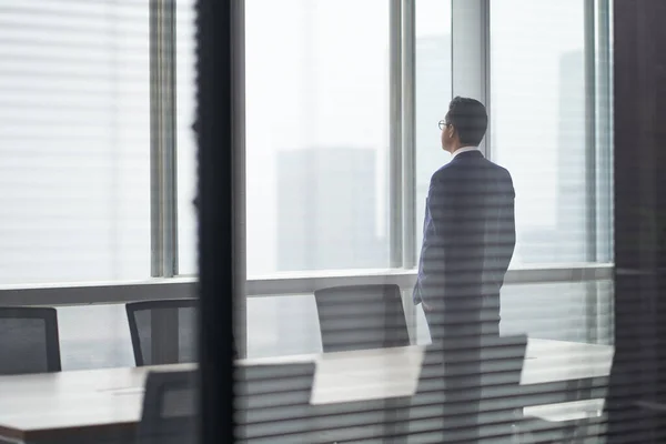 rear side view of an asian business man standing in front of office window looking out and thinking hands in pockets