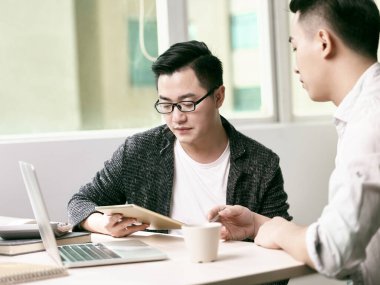 İki genç Asyalı girişimci dijital tablet ve dizüstü bilgisayar kullanarak ofiste iş konuşuyorlar