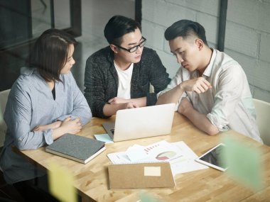 Ofiste birlikte çalışan üç Asyalı genç girişimci iş planını dizüstü bilgisayarla tartışıyor.