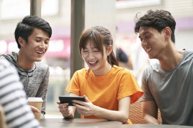 Üç genç Asyalı yetişkin birlikte cep telefonu kullanarak açık kahve dükkanında oturuyor.